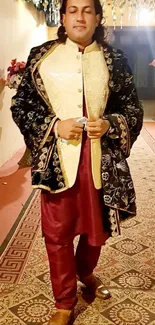 Man in elegant traditional attire with ornate jacket and burgundy robe on patterned rug.