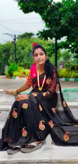 Woman wearing black saree sitting outdoors.