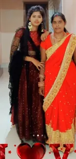 Two women in elegant sarees, one red, standing indoors.