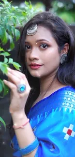 Woman in blue traditional attire, adorned with jewelry, surrounded by greenery.