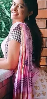 Woman in pink patterned sari smiling by a brick wall with greenery.