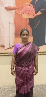 A woman in purple traditional attire stands elegantly in front of a framed picture.