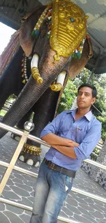 A man in blue shirt standing beside a large decorated elephant statue outdoors.