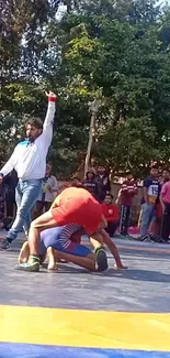 Outdoor wrestling match with referee and spectators.