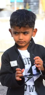 Young child in black blazer with sunglasses in urban background.