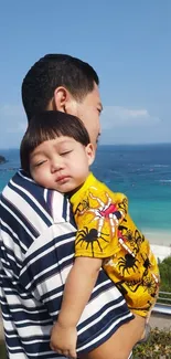 Father holding sleeping child by the beach with ocean background.