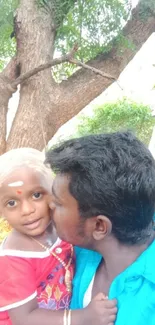 Father and child embracing under a large tree with lush greenery.