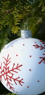 White Christmas ornament with red snowflake patterns hanging on green pine branches.