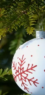 White ornament with red snowflakes hanging on evergreen branch.