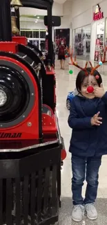 Child with antlers beside red and black train in a mall setting.