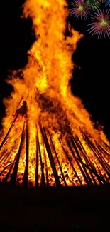 Towering bonfire with colorful fireworks in night sky.