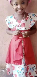 Smiling girl in floral dress with red sash, standing against a pink textured wall.