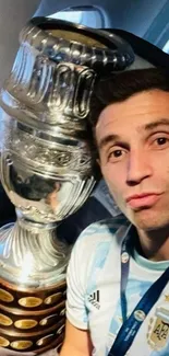 Footballer posing with silver trophy in bright setting.