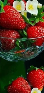 Bright strawberries with flowers in a clear glass bowl on a dark background.