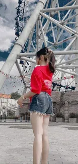 Girl stands near Ferris wheel wearing red shirt and denim shorts.