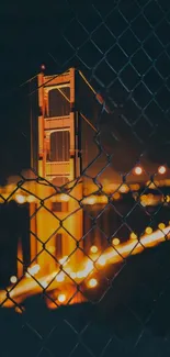 Golden Bridge at night through a chain-link fence with glowing city lights.