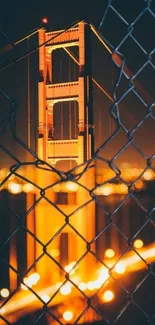 Golden Gate Bridge at night with orange glow through a chain-link fence.