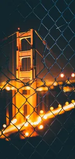 Golden Gate Bridge at night viewed through a chain fence, with warm orange lighting.