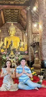 Golden Buddha statue in an ornate temple, with two people in prayer pose.