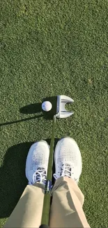 Golf ball and putter on green grass with shoes visible.