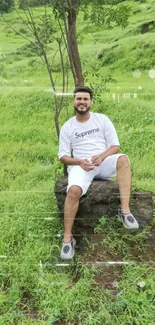 Man sitting on a rock in a lush green field with trees and hills in the background.