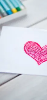 Pink crayon-drawn heart on a white paper background with pencils.