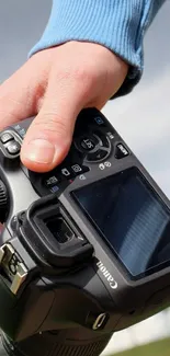 Hand gripping a DSLR camera against a cloudy sky backdrop.