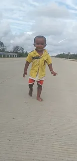 Child in yellow shirt walking on a gray road under a cloudy sky.