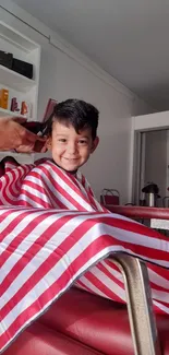 A happy child getting a haircut in a red-striped barber cape.