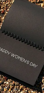 Elegant black notebook with Happy Women's Day message on pebbles.