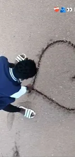 A person drawing a heart in the sand on a beach, wearing a blue shirt.