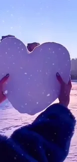 Hands hold a heart-shaped snow against a city skyline at evening.