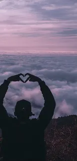 A person forms a heart silhouette against a purple evening sky over clouds and mountains.