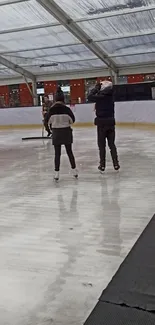 Couple ice skating indoors with a clear roof and warm ambient lights.