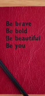 Red notebook with motivational quotes on a wooden desk, alongside a pencil and coffee cup.