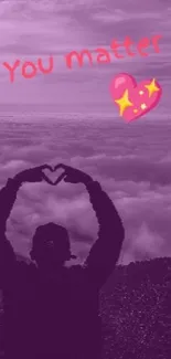 Silhouette forming heart with hands over a purple mountain landscape.