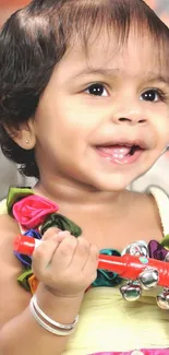 Smiling baby holding a red toy, dressed in colorful outfit.