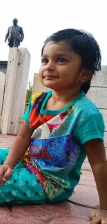 A joyful child in colorful clothes sitting on a paved ground outdoors.