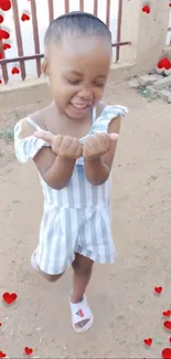 Smiling child in striped outfit surrounded by red hearts outdoors.