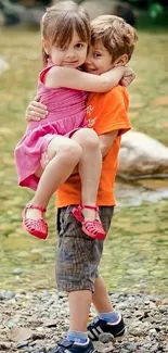 Two children joyfully embrace by a forest stream.