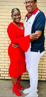 Couple embracing happily in red outfit against brick wall.