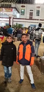 Two children in coats pose happily at an outdoor evening gathering.