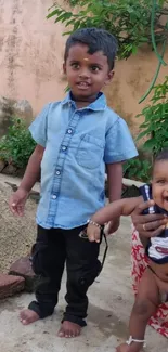 Two children smiling outdoors with a beige wall background.