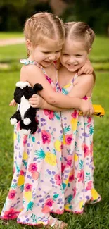 Two young twins in floral dresses hug in a lush green garden.