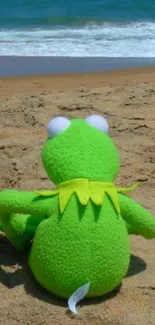 Green frog toy sitting on the sandy beach with ocean waves in the background.