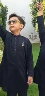 Two kids in black traditional outfits raising hands in a green garden.