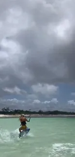 Kiteboarder gliding on turquoise water under a cloudy sky.