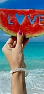 Hand holding watermelon with 'LOVE' by beach.