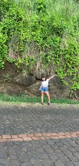 Girl stands by a lush, green wall on a cobblestone street.