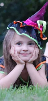 Child in a colorful witch costume lying on grass.